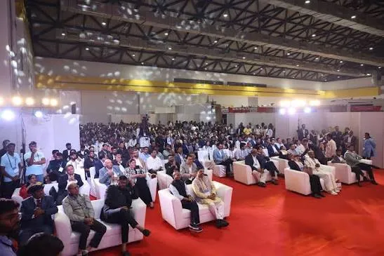 A wide conference hall with a lit stage, carpeted aisle and a full seated audience.