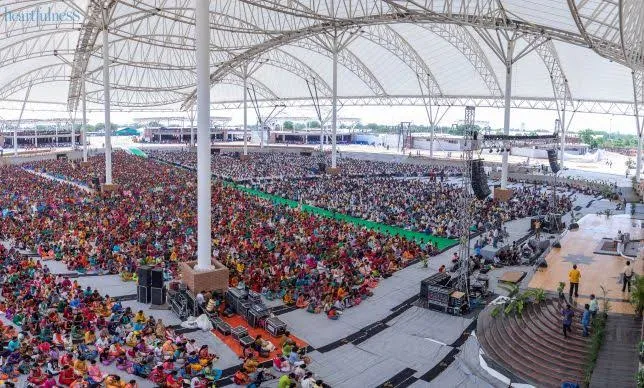 Aerial view over an immense clear-span canopy sheltering a seated assembly of many thousands.